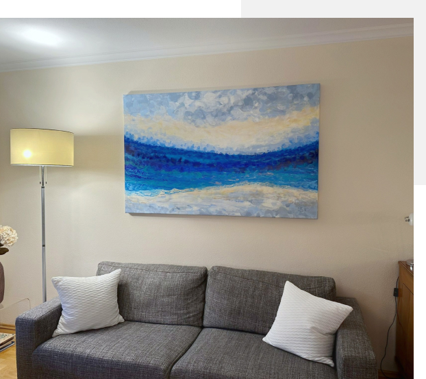 Painting in blue, white and gray hanging on a pale yellow wall behind a gray sofa with white pillows.